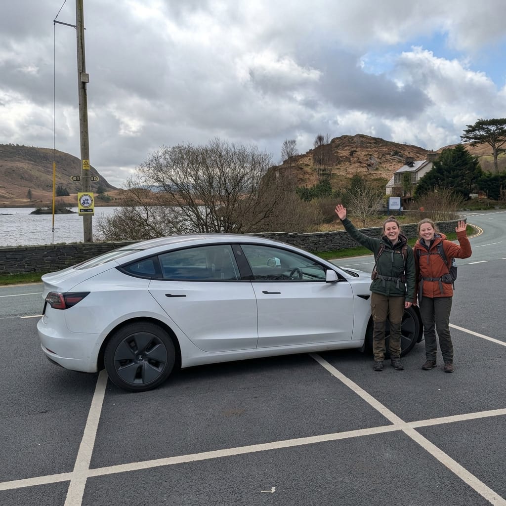 Two happy hikers waving beside a white Tesla Model 3 taxi in a scenic North Wales car park, with a lake and hills in the background.