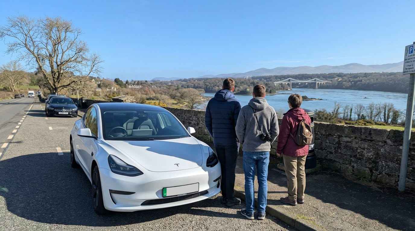 White Tesla Model 3 taxi parked at Menai Strait viewpoint while passengers enjoy a sunny March view of the Menai Suspension Bridge and Snowdonia mountains in North Wales.